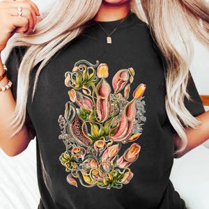 Puede incluir: Camiseta negra con una ilustración botánica de plantas carnívoras en tonos rosa, amarillo y verde. El diseño presenta arte botánico detallado, ideal para amantes de las plantas y ropa original.