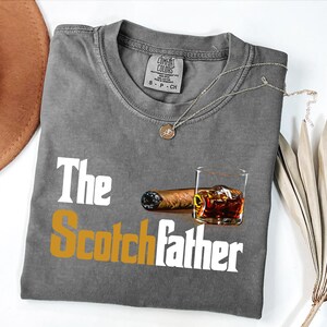 a gray t-shirt with the text "The Scotchfather" printed on it, along with an image of a cigar and a glass of whiskey.