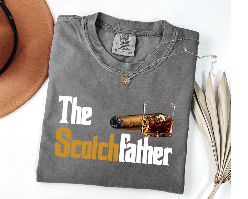 a gray t-shirt with the text "The Scotchfather" printed on it, along with an image of a cigar and a glass of whiskey.