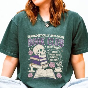A person wearing a green t-shirt with a graphic design featuring a skull, books, and other symbols, along with text that reads "Unaopoligically anti-social book club meets weekly making friends with characters, not people".