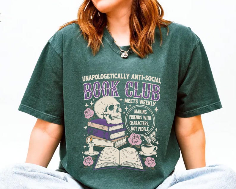 A person wearing a green t-shirt with a graphic design featuring a skull, books, and other symbols, along with text that reads "Unaopoligically anti-social book club meets weekly making friends with characters, not people".