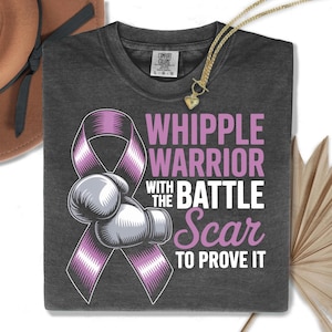 Puede incluir: Camiseta gris oscuro con el texto "WHIPPLE WARRIOR WITH THE BATTLE SCAR TO PROVE IT" en blanco y rosa. El diseño incluye una cinta morada y guantes de boxeo. Un collar dorado con un colgante de corazón está sobre la camiseta.