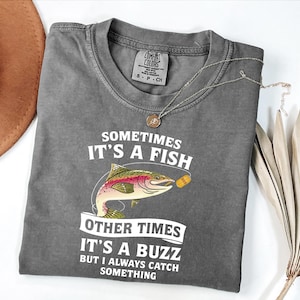 Könnte beinhalten: Ein graues Comfort Colors T-Shirt mit dem Text "Sometimes It's A Fish, Other Times It's A Buzz, But I Always Catch Something" und einer Grafik einer Regenbogenforelle. Eine silberne Halskette mit Anhänger befindet sich auf dem Shirt.