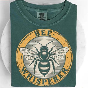 May include: A dark teal t-shirt with a circular graphic featuring a detailed illustration of a bee. The bee is centered within a yellow ring, and the words "BEE WHISPERER" are printed around the bee in a vintage style font. The shirt is folded.