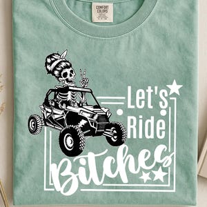 May include: A light green Comfort Colors t-shirt featuring a black and white graphic of a skeleton driving an off-road vehicle. The text "Let's Ride Bitches" is displayed in white script. The shirt has a crew neck and a tag.