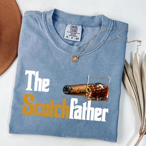A blue t-shirt with the text "The Scotchfather" printed on it, along with an image of a cigar and a glass of whiskey.