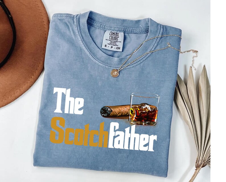 A blue t-shirt with the text "The Scotchfather" printed on it, along with an image of a cigar and a glass of whiskey.