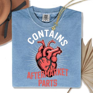 Könnte beinhalten: Hellblaues T-Shirt mit dem Text "CONTAINS AFTERMARKET PARTS" über und unter einer roten anatomischen Herzillustration. Eine goldene Halskette mit Herzanhänger liegt auf dem Shirt. Das Shirt ist von der Marke Comfort Colors.