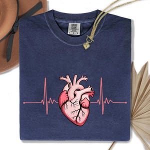 Könnte beinhalten: Marineblaues T-Shirt mit einem rosa anatomischen Herz und einer rosa Herzschlaglinie. Eine goldene Halskette mit Herzanhänger liegt auf dem T-Shirt. Das T-Shirt hat einen Rundhalsausschnitt und kurze Ärmel. Der Markenname "Comfort Colors" ist auf dem Etikett aufgedruckt.