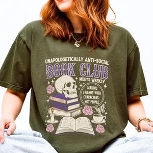 A person wearing a green t-shirt with a graphic design that says "Unaopoligically anti-social book club meets weekly" and includes a skull, books, and other symbols.