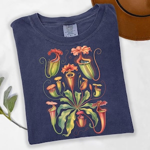 May include: Navy blue t-shirt with a botanical illustration of pitcher plants. The design showcases various pitcher plant species in green, orange, and red hues. The shirt is made of a soft material and has the Comfort Colors brand tag.