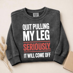 Puede incluir: Sudadera gris oscuro con el texto "QUIT PULLING MY LEG SERIOUSLY. IT WILL COME OFF" en letras blancas y rojas. La sudadera es de la marca Comfort Colors.