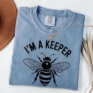 May include: A light blue t-shirt with the text "I'M A KEEPER" above a black and white bee illustration. The shirt has a crew neck and short sleeves. A gold necklace with a pendant is draped on the shirt.