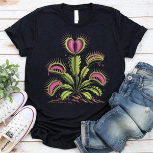 Puede incluir: Camiseta negra con un diseño colorido de una planta carnívora Venus atrapamoscas. El diseño incluye elementos rosas y verdes, con una flor en forma de corazón. Se muestra con vaqueros azules y zapatillas blancas.