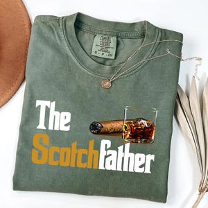 a green t-shirt with a graphic design depicting a glass of whiskey and a cigar, along with the text "The Scotchfather".