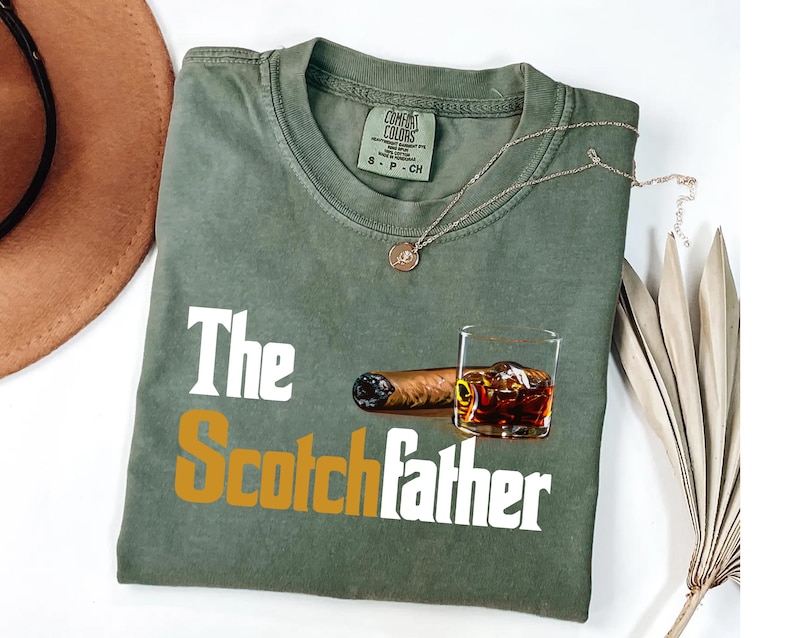 a green t-shirt with a graphic design depicting a glass of whiskey and a cigar, along with the text "The Scotchfather".