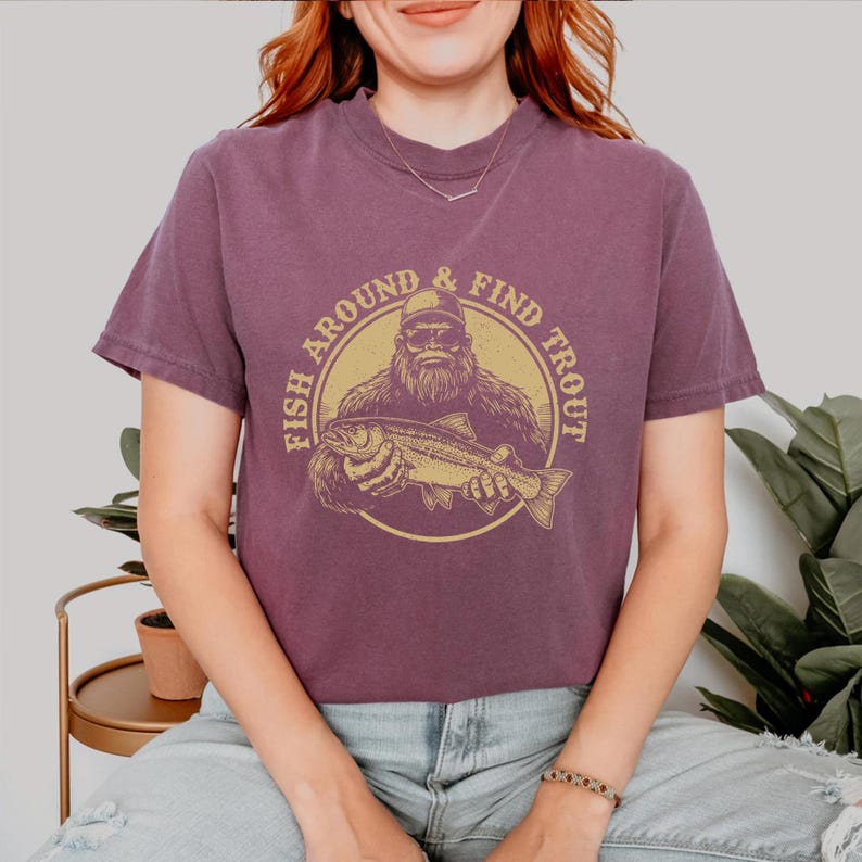 A woman wearing a purple t-shirt with a graphic of a bearded fish and the text "FISH AROUND & FIND TROUT" printed on it.