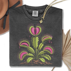 May include: Dark gray Comfort Colors t-shirt featuring a Venus flytrap design with pink and green details. The graphic includes a heart-shaped flower and several open traps. A gold necklace with a heart pendant is draped over the shirt.