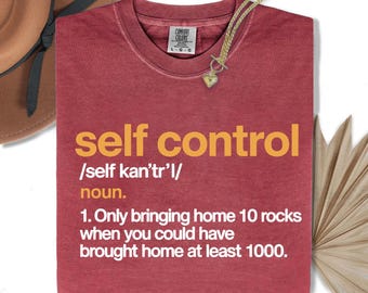 Op de afbeelding: Een bordeauxrood t-shirt met de gele woorden "self control" en een definitie. Een gouden ketting met een hartvormige hanger ligt op het shirt. Een bruine hoed is linksboven zichtbaar.