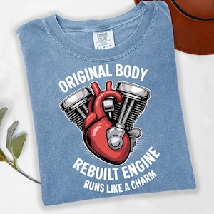 May include: Light blue t-shirt with a graphic of a red heart and engine parts. The text reads "ORIGINAL BODY REBUILT ENGINE RUNS LIKE A CHARM". The shirt has a crew neck and short sleeves.