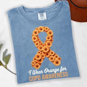 Puede incluir: Camiseta azul claro con un diseño de lazo de girasoles naranjas para la concienciación sobre la EPOC. El lazo está hecho de girasoles naranjas y el texto "I Wear Orange for COPD Awareness" está impreso debajo.