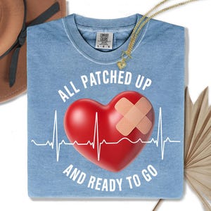 Könnte beinhalten: Hellblaues Comfort Colors T-Shirt mit dem Text "ALL PATCHED UP AND READY TO GO". Die Grafik zeigt ein rotes Herz mit einem Pflaster und einer weißen EKG-Linie. Eine goldene Halskette mit Herzanhänger ist sichtbar.