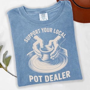 Könnte beinhalten: Hellblaues T-Shirt mit dem weißen Schriftzug "SUPPORT YOUR LOCAL POT DEALER". Die Grafik zeigt Hände, die einen Topf auf einer Töpferscheibe formen. Das Shirt besteht aus einem weichen Material und hat einen Rundhalsausschnitt.