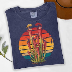 May include: A navy blue t-shirt featuring a graphic of pitcher plants in front of a retro sunset design. The plants are red, yellow, and green. The shirt is made by Comfort Colors.