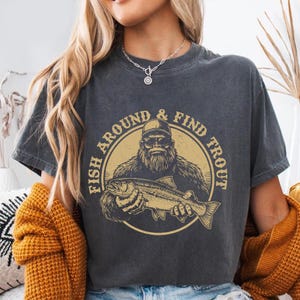 A woman wearing a gray t-shirt with a graphic design of a bearded man holding a fish, along with the text "FISH AROUND & FIND TROUT".