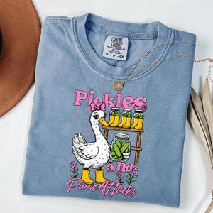May include: A blue short-sleeve t-shirt featuring a graphic of a white goose wearing a pink bow and yellow boots. The goose stands near a wooden shelf holding jars of pickles. The text reads Pickles and Panic Attacks in pink lettering.