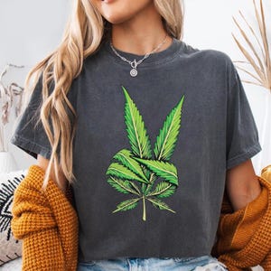 May include: Dark gray t-shirt featuring a vibrant green cannabis leaf design. The graphic is detailed with various shades of green, creating a visually appealing contrast against the shirt's color. The shirt has short sleeves and a crew neck.