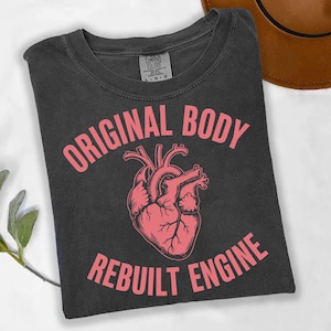May include: Dark gray t-shirt with the words "ORIGINAL BODY" above a pink anatomical heart illustration and the words "REBUILT ENGINE" below. The shirt is folded and has a Comfort Colors tag.