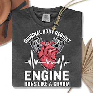 May include: Dark gray t-shirt with the text "ORIGINAL BODY REBUILT ENGINE RUNS LIKE A CHARM". The graphic includes a red heart, pistons, and a heartbeat line. A gold necklace with a heart pendant is draped over the shirt.