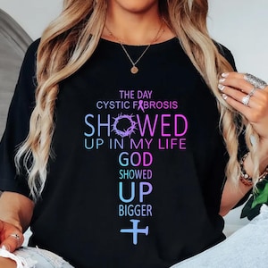 Puede incluir: Camiseta negra con un gráfico colorido que dice: "The day Cystic Fibrosis showed up in my life, God showed up bigger." El diseño incluye una corona de espinas, una cinta y una cruz.