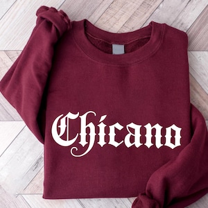 Könnte beinhalten: Ein weinrotes Sweatshirt mit dem Wort "Chicano" in weißer, gotischer Schrift auf der Vorderseite. Das langärmlige Oberteil ist auf einer hellen Holzmaserung gefaltet.