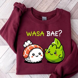 Puede incluir: Sudadera burdeos con el texto "WASA BAE?" en verde y blanco. El diseño presenta un rollo de sushi de dibujos animados y un personaje de wasabi con caras sonrientes. La sudadera está hecha de un material suave.