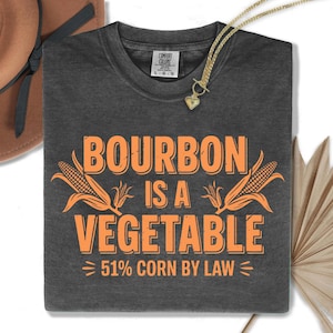 Puede incluir: Camiseta gris oscuro con el texto naranja "BOURBON IS A VEGETABLE" y dibujos de maíz. También incluye el texto "51% CORN BY LAW". Se ve un collar dorado y un sombrero marrón.