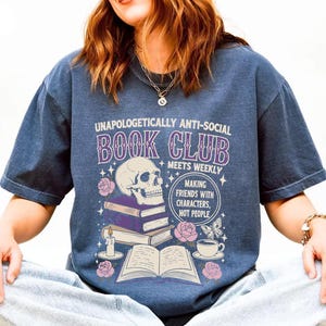 A person wearing a blue t-shirt with a graphic design featuring a skull, books, and other symbols, along with text that reads "Unauthorically anti-social book club meets weekly making friends with characters not people".