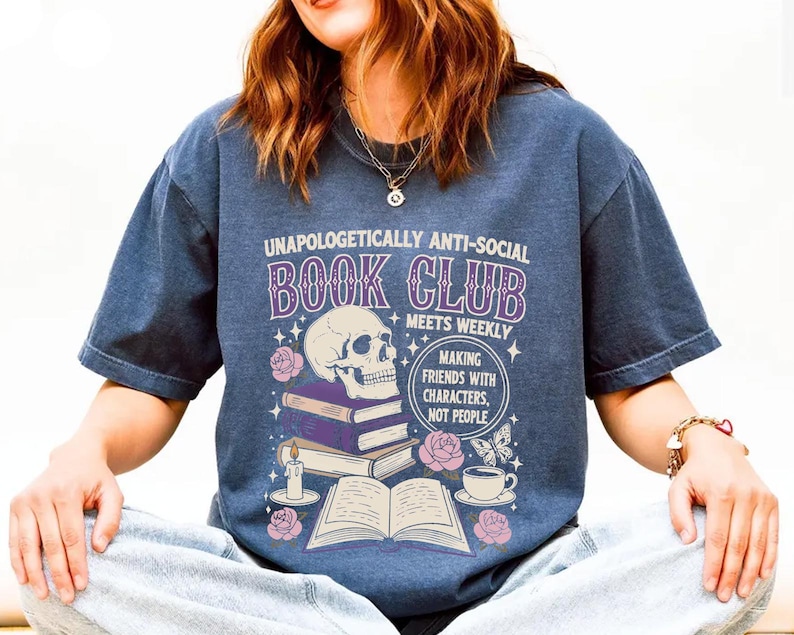 A person wearing a blue t-shirt with a graphic design featuring a skull, books, and other symbols, along with text that reads "Unauthorically anti-social book club meets weekly making friends with characters not people".