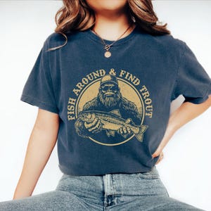 A woman wearing a blue t-shirt with a graphic design of a bearded fish and the text "FISH AROUND & FIND TROUT" printed on it.