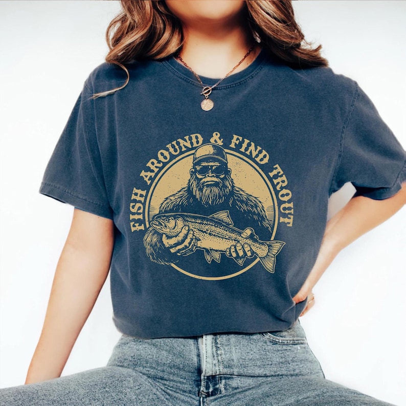 A woman wearing a blue t-shirt with a graphic design of a bearded fish and the text "FISH AROUND & FIND TROUT" printed on it.