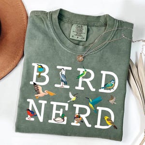 May include: Olive green t-shirt with the words "BIRD NERD" in white block letters. The shirt features colorful illustrations of various birds. A silver necklace with a small pendant is draped over the shirt.