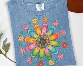 May include: A light blue t-shirt with a large, colorful flower design. The flower has rainbow-colored petals and a detailed center. Smaller flowers and green circles surround the main design. The shirt has a crew neck and a tag that says "COMFORT COLORS".