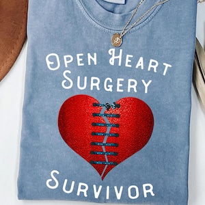 May include: A blue t-shirt with the words "Open Heart Surgery Survivor" in white. A red heart graphic is stitched together with blue thread. A gold necklace with a pendant is draped on the shirt.