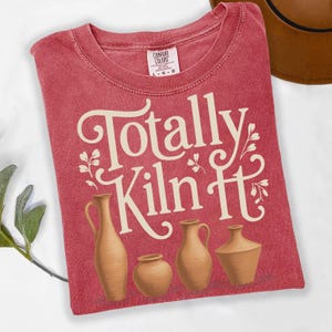 Puede incluir: Camiseta roja con el texto "Totally Kiln It" en una fuente decorativa. Debajo del texto hay ilustraciones de cuatro vasijas de cerámica marrones. La camiseta tiene cuello redondo y una etiqueta Comfort Colors.