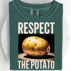 May include: A dark teal t-shirt with the words "RESPECT THE POTATO" in white block letters. A close-up image of a potato is centered below the word "RESPECT". The potato is yellow with brown spots.