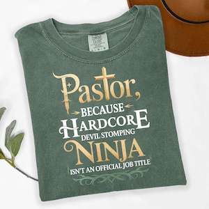 Könnte beinhalten: Ein salbeigrünes T-Shirt mit dem Text "Pastor, because Hardcore Devil Stomping Ninja Isn't An Official Job Title" in gold- und weißfarbenen Buchstaben. Das Shirt hat einen Rundhalsausschnitt und ein Comfort Colors-Etikett.