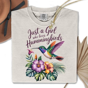 May include: A light beige Comfort Colors t-shirt featuring a colorful hummingbird design with the text "Just a Girl who loves Hummingbirds." The design includes flowers and tropical leaves. A gold necklace is draped over the shirt.