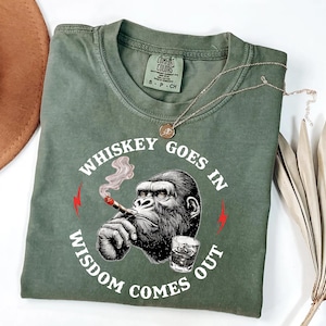 May include: Green t-shirt with a graphic of a gorilla smoking a cigar and a glass of whiskey. The text reads "Whiskey Goes In, Wisdom Comes Out". The shirt has a crew neck and short sleeves.