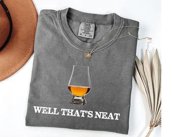 May include: A gray Comfort Colors t-shirt with the text "WELL THAT'S NEAT" in white. The shirt features a graphic of a whiskey glass filled with amber liquid. A silver necklace with a pendant is on the shirt.